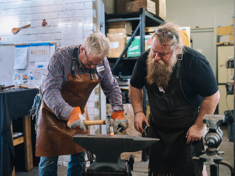 Two men with tools standing over an anvil at a blacksmithing workshop